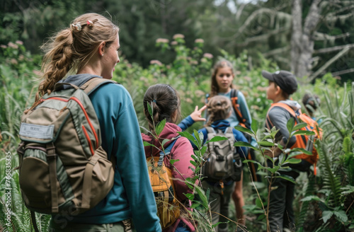 A teacher and group of children were on an outdoor field trip in nature, learning about plants from the female naturalist smiling at the camera, standing among tall grasses with backpacks beside them