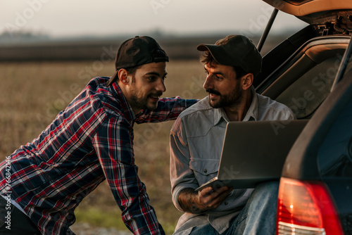 Two men in trendy attire utilizing a laptop from the back of a car.
