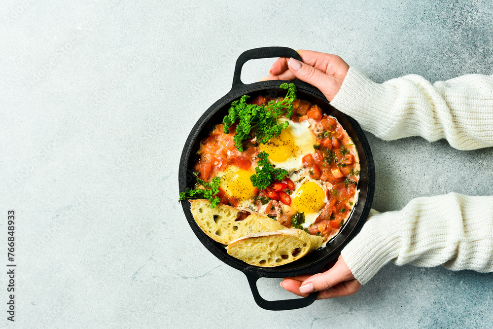 Hands hold a frying pan with Shakshuka. Poached eggs in a spicy tomato