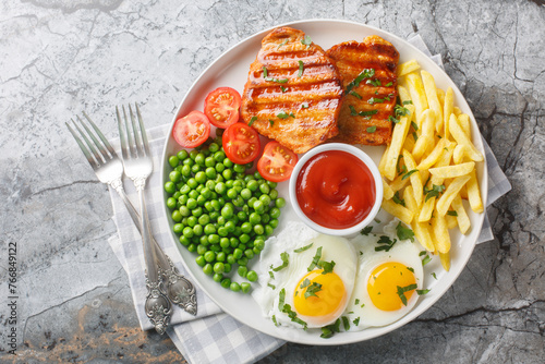 Tender grilled steak served with crisp golden French fries, fied eggs, green pea and fresh tomato closeup on the plate on the table. Horizontal top view from above © FomaA