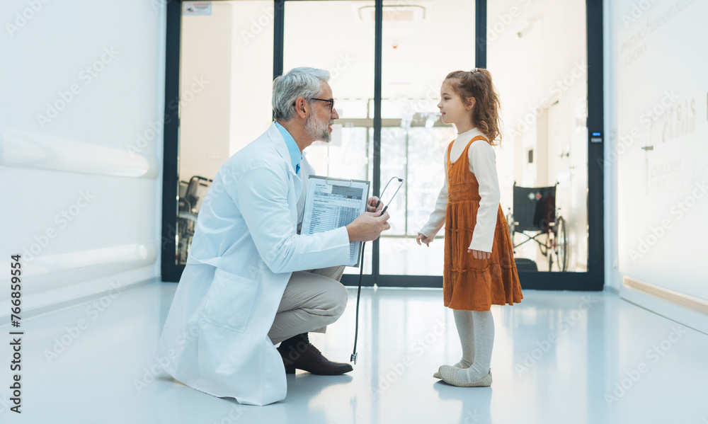 Obraz premium Little girl talking with doctor in hospital lobby. Friendly relationship between helathcare worker and child patient.