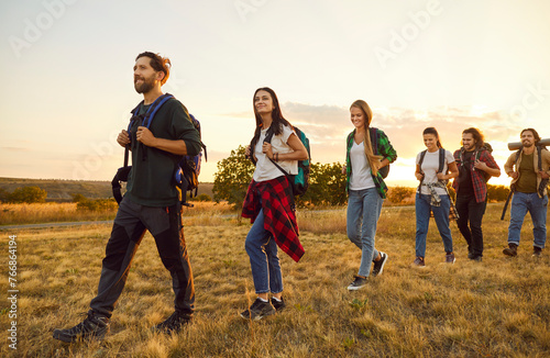 Group of young friends tourists trekking with backpacks walking in a row in the field. Happy people men and women on a hiking trip holiday in nature at sunset. Adventure, travel and tourism concept.