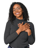 African American woman in studio setting has friendly expression, pressing palm to chest. Love concept.