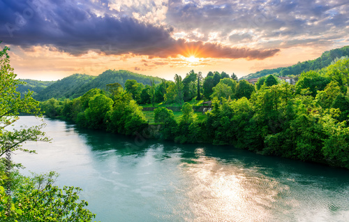 beautiful landscape of spring or summer sunset river with blue water and green hills on sides and mountains with amazing cloude sky on background