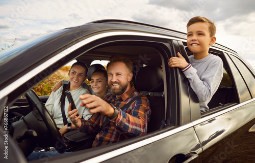 Portrait of happy smiling family of four with kids sitting inside car driving automobile. Father using digital map application and showing way or pointing to a landmark outdoors. Road trip concept.