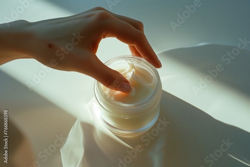 Female hand dipping fingers into a jar of cosmetic cream, soft light and focus, gentle colors. Concept of self-care, skin care products, pampering and relaxation in skincare routine.