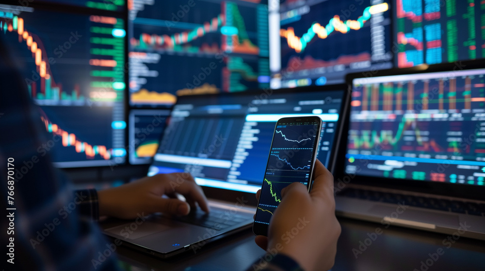Fototapeta premium person is engaged in monitoring financial data. They are using both a smartphone and a laptop, with additional larger screens in the background