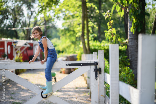 Beautiful young girl sitting on white wooden fence, helping on family farm during summer.