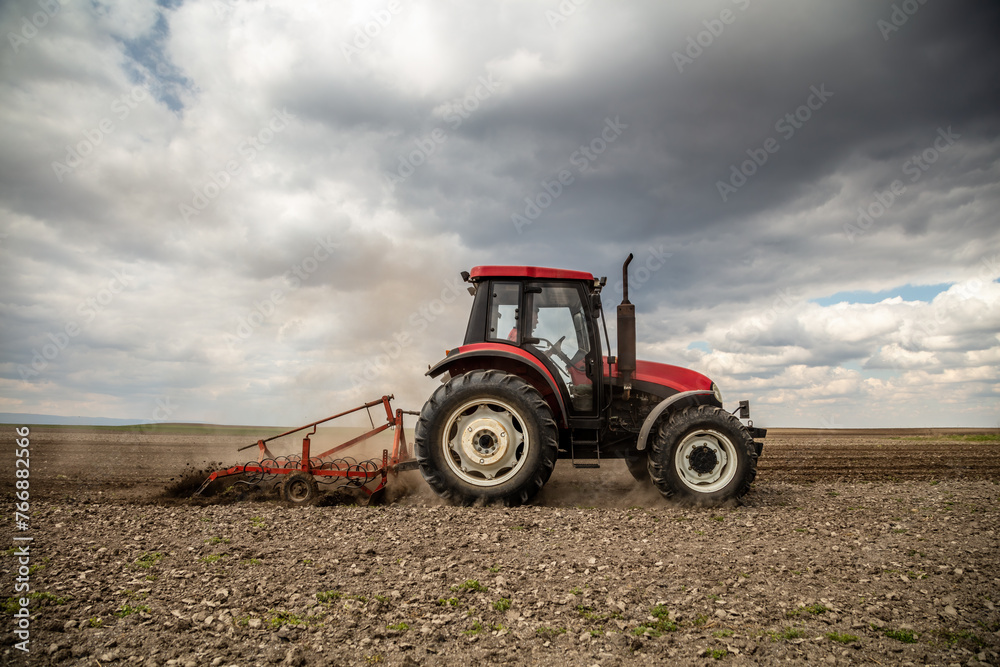 Obraz premium Powerful red tractor tills the soil on a vast farm with a dramatic cloudy sky overhead