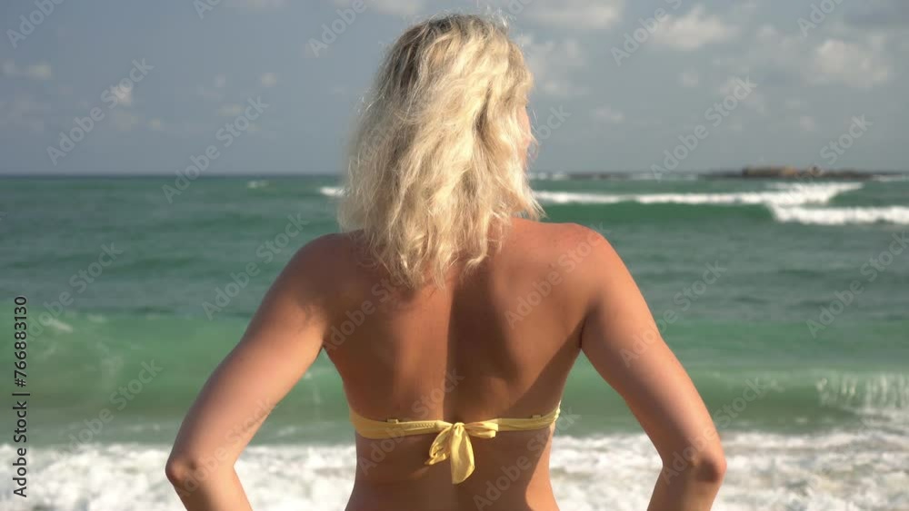 A young attractive blonde woman in a swimsuit stands with her back against the background of the sea. Tropical sea beach holiday A young attractive blonde woman in a swimsuit stands with her back against the background of the sea. Tropical sea beach holiday