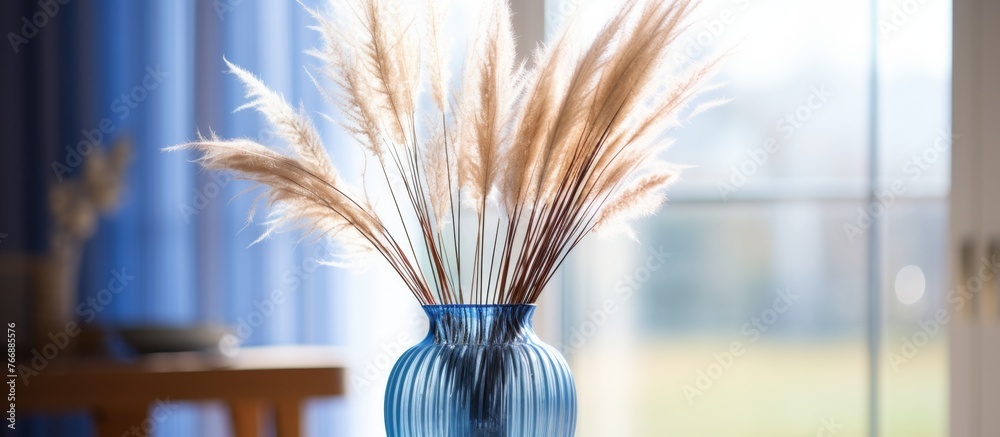 Terrestrial plants in electric blue vase on hardwood table, complemented by staple food of dried flowers. Still life photography art capturing the serene event with grass outside the window - obrazy, fototapety, plakaty 