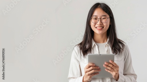Standing over a white background, an Asian woman in glasses holds a tablet and smiles, wearing glasses.