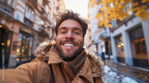 Handsome young man in casual clothing smiling while taking selfie outdoors. Caucasian tourist talking with friends by smartphone while walking on city streets. Vlogging photo