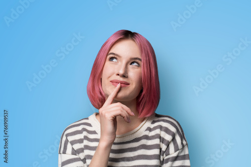 Portrait ad image - mysteriously grinning smile look up thinking gladly pink hair woman wear grey striped sweater jumper, hold finger over her she lips, isolated blue studio wall background.
