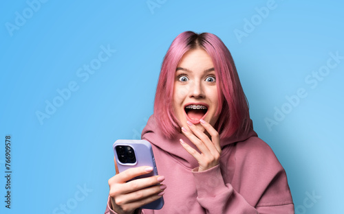 Shocked astonished very happy pink woman wear braces, hoodie open mouth, using holding hand cell phone, modern smartphone, mobile phone, isolated blue wall background. Online offer, dental care ad.