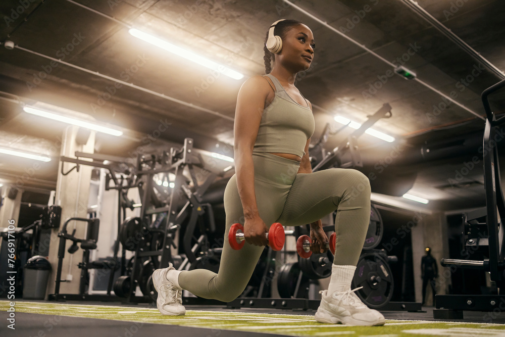 Naklejka premium A fit young black sportswoman practicing lunges with dumbbells and listening music on headphones at gym.