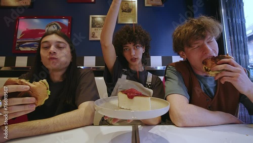 The frame captures three friends in a diner, presumably in their twenties. A male with long dark hair, clad in a black shirt, voraciously bites into a burger, savoring every taste. Centered is a young
