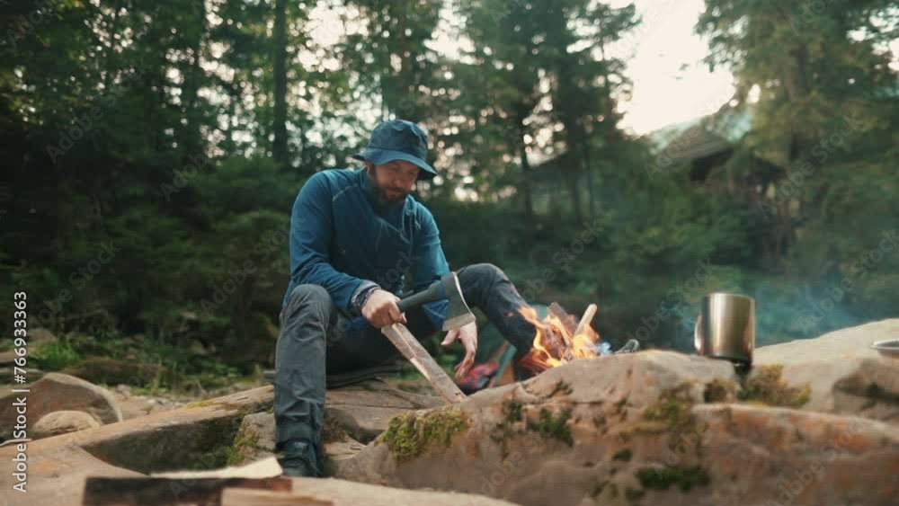 Close-up of a lone tourist sitting on a stone in a tourist camp near a fire and chopping wood Close-up of a lone tourist sitting on a stone in a tourist camp near a fire and chopping wood