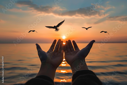 Hands outstretched upwards in a gesture of worship against a serene sunset over water, with birds soaring above. Symbolizing prayer and seeking divine blessings