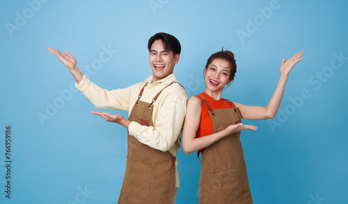 Happy young asian barista couple partnership wearing apron standing and open hand welcome isolated on blue background.