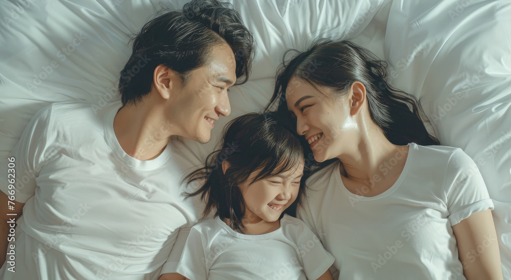 A young Chinese couple and their daughter lay on the bed, smiling at each other, wearing white T-shirts