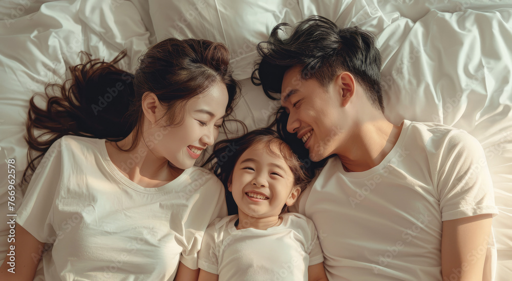 A young Chinese couple and their daughter lay on the bed, smiling at each other, wearing white T-shirts