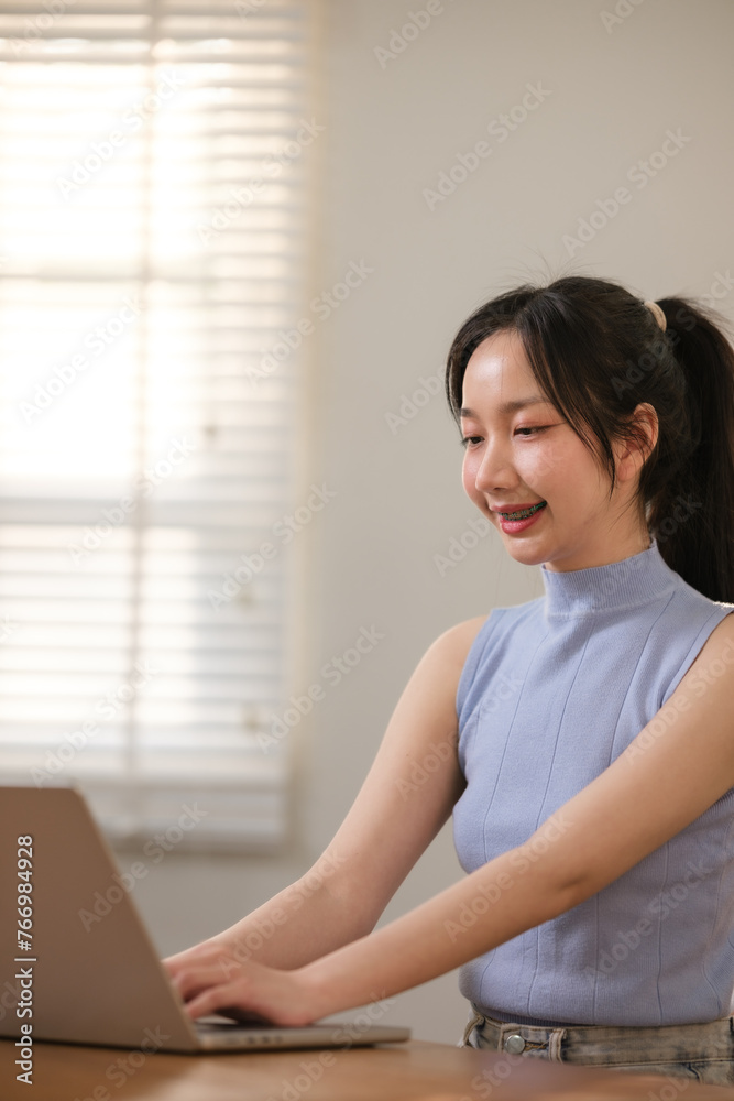 Young attractive happy Asian female student sitting in the living room smiling and looking at the camera while working on a laptop at the home office. Young attractive happy Asian female student sitting in the living room smiling and looking at the camera while working on a laptop at the home office.