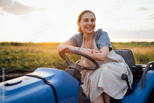 A young smiling farmer sits at the wheel of a tractor. A funny charming woman in casual clothes cultivates an agricultural field. © Dasha Petrenko