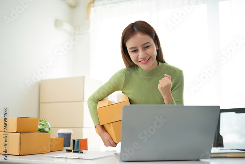 Startup SME small business entrepreneur of freelance Asian woman using a laptop with box Cheerful success Asian woman her hand lifts up online marketing packaging box and delivery SME idea concept © chartchai
