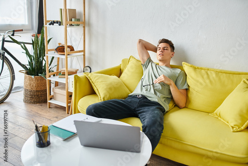 weary guy with laptop putting hand behind head and leaning on yellow couch with glasses © LIGHTFIELD STUDIOS