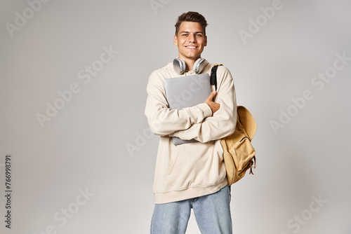 cheerful student in 20s with laptop and headphones confident standing against grey background