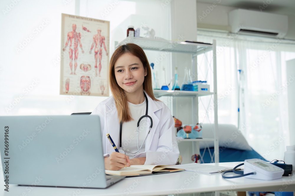 Young Asian doctor in white medical uniform with stethoscope using laptop computer video chatting with patient at table in health clinic or hospital Young Asian doctor in white medical uniform with stethoscope using laptop computer video chatting with patient at table in health clinic or hospital