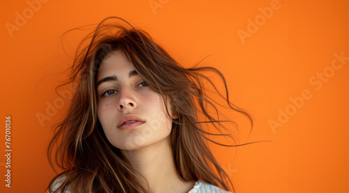 A woman with long hair is standing in front of an orange wall. She has a serious expression on her face