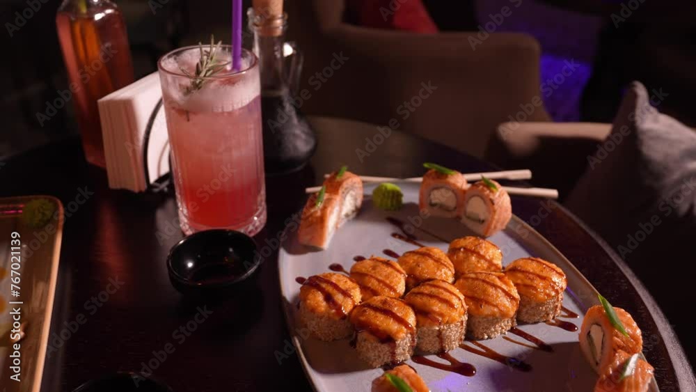 Set of various delicious mouth-watering sushi rolls on the table with a cocktail, soy sauce, chopsticks and a vase with a flower in the restaurant in the evening, a selective focus. Set of various delicious mouth-watering sushi rolls on the table with a cocktail, soy sauce, chopsticks and a vase with a flower in the restaurant in the evening, a selective focus.
