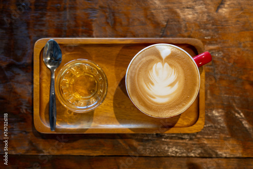 High-angle view of hot coffee in glass decorated with milk foam. Served with syrup in a clear glass for those who like sweetness. Placed on a wooden tray and served on a table for a classic look.