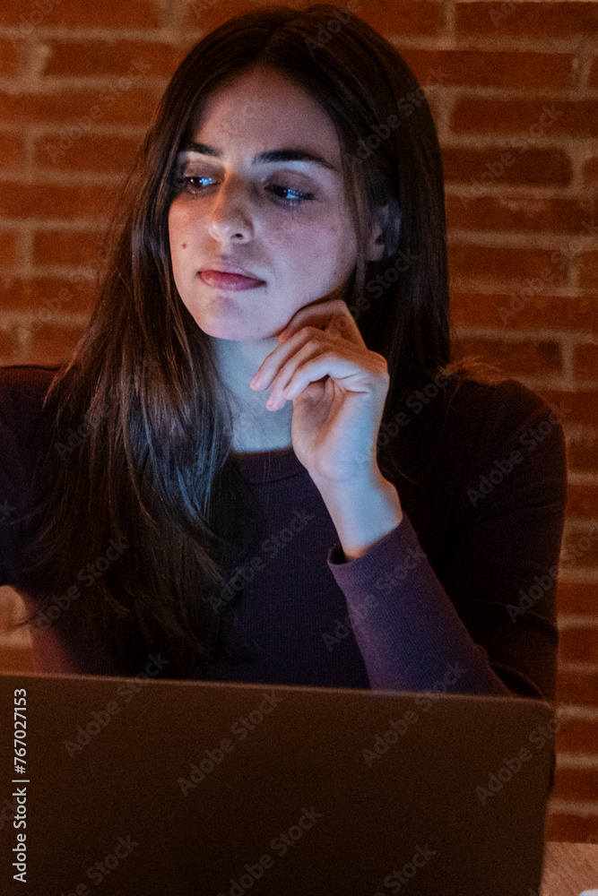 Young pretty smiling woman using laptop in home at night. Sad girl looking at computer. Cheerful people sitting at desk in living room. Concept of enterprising, telecommuting and hardworking person. Young pretty smiling woman using laptop in home at night. Sad girl looking at computer. Cheerful people sitting at desk in living room. Concept of enterprising, telecommuting and hardworking person.