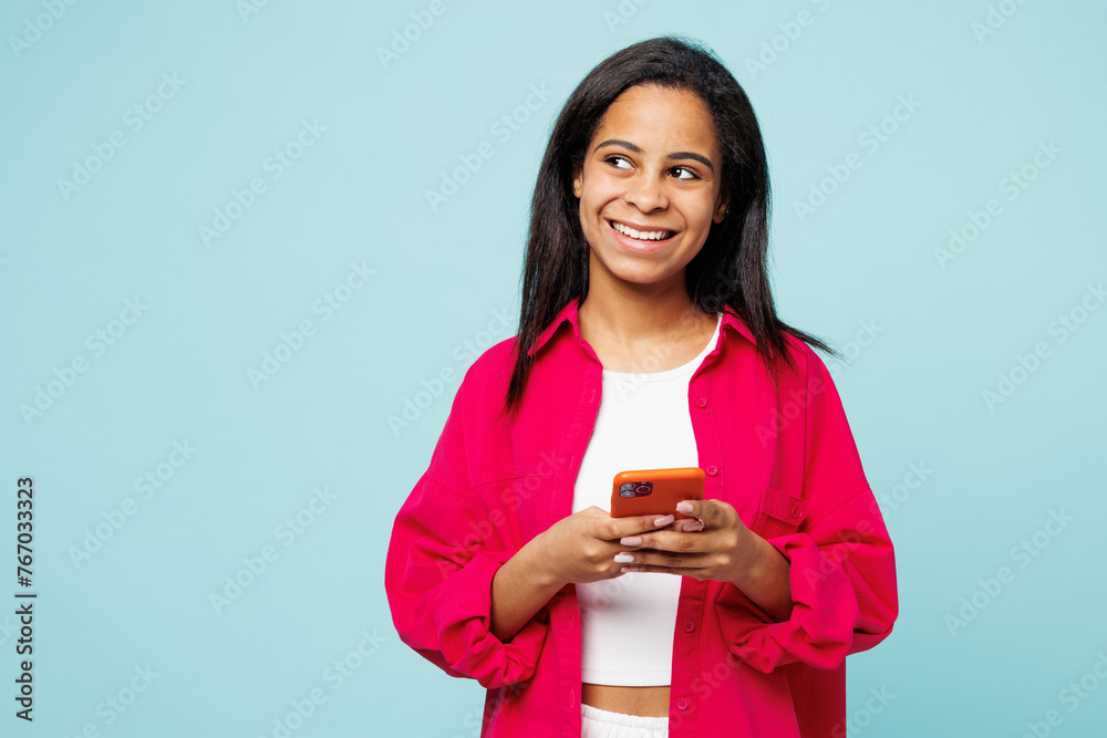 Little kid teen girl of African American ethnicity wears pink shirt white t-shirt hold in hand use mobile cell phone look aside isolated on plain pastel blue background. Childhood lifestyle concept. Little kid teen girl of African American ethnicity wears pink shirt white t-shirt hold in hand use mobile cell phone look aside isolated on plain pastel blue background. Childhood lifestyle concept.
