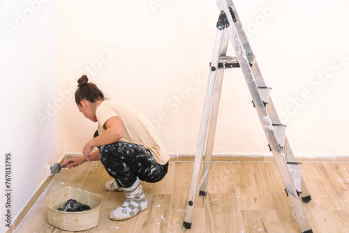 A woman is diligently painting the corner of a room, with paint supplies and a ladder nearby, showing meticulous DIY home improvement