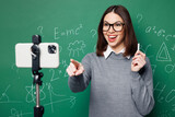 Young smart teacher woman wear grey casual shirt glasses talk speak use camera of mobile cell phone isolated on green wall chalk blackboard background studio Education in high school college concept