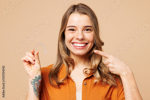 Young happy woman she wear orange shirt casual clothes hold in hand invisible transparent aligners, invisalign bracer point on teeth isolated on plain pastel light beige background. Lifestyle concept. photo