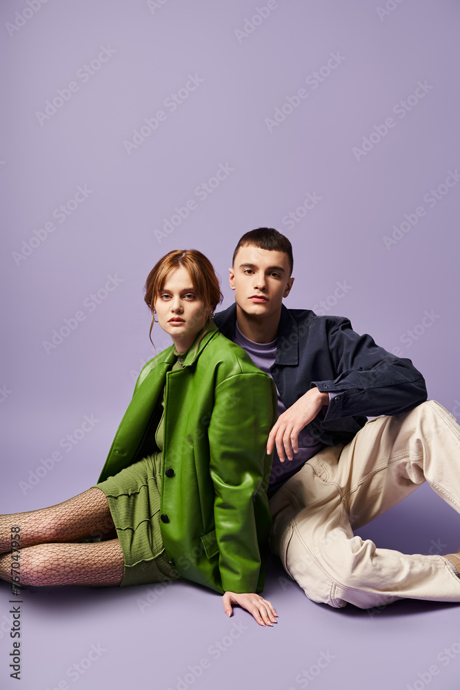 Obraz premium fashionable couple in vibrant attires sitting on floor and looking at camera on purple backdrop