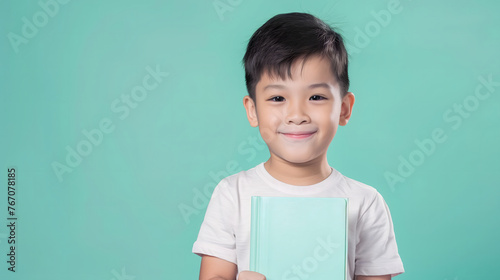 A happy and smiling Asian boy is holding a blue book on a plain green background with copy-space for text.