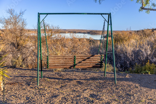 Metal framed wooden swinging bench attached with chains, on gravel overlooking a body of water and bushes in the distance. 