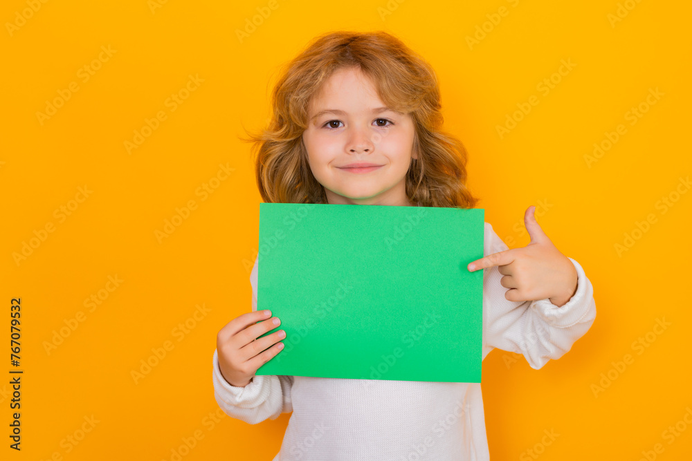 Smiling kid showing index finger on green empty sheet of paper, isolated on yellow background. Portrait of a kid holding a blank placard, poster. Smiling kid showing index finger on green empty sheet of paper, isolated on yellow background. Portrait of a kid holding a blank placard, poster.