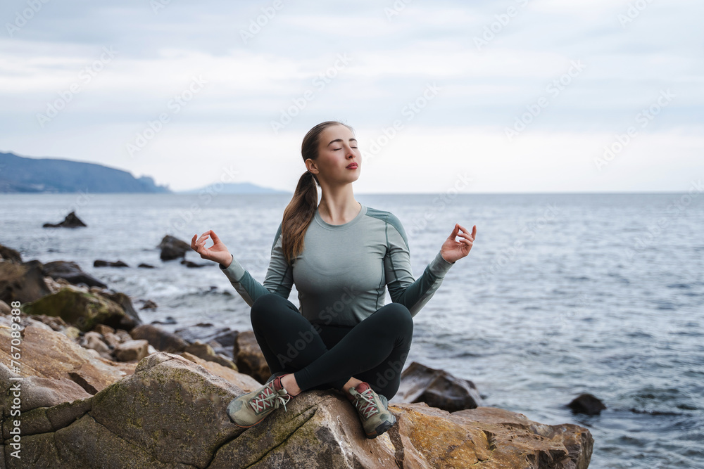 Young spiritual calm happy woman with closed eyes meditate in lotus pose at the sea, om aum gesture, girl relaxing outdoors, meditation, try to calm down in the mountains, female doing yoga asana Young spiritual calm happy woman with closed eyes meditate in lotus pose at the sea, om aum gesture, girl relaxing outdoors, meditation, try to calm down in the mountains, female doing yoga asana