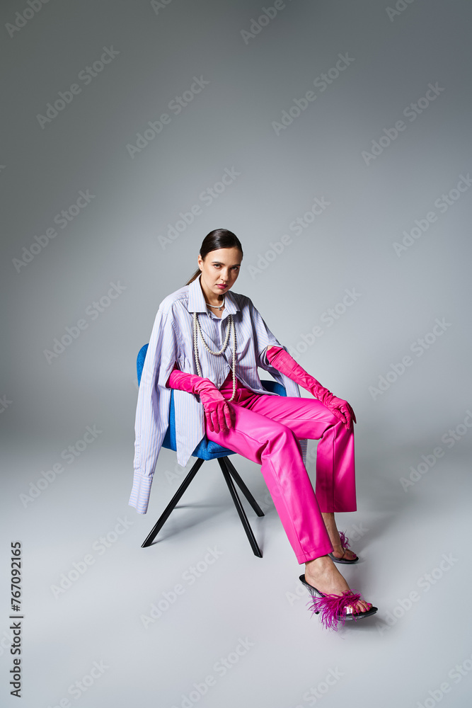 Chic brunette girl in stylish pink outfit touching her legs, sitting on chair on grey background Chic brunette girl in stylish pink outfit touching her legs, sitting on chair on grey background