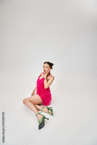 Playful girl in trendy pink dress and green shoes touching her cheek and sitting on grey background