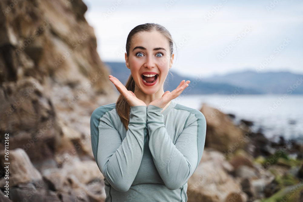 Young woman looking happy and excited, shocked with an unexpected surprise with both hands open next to face, sporty girl with bulging raise hands and shouts, exclaims in amazement, expresses shock