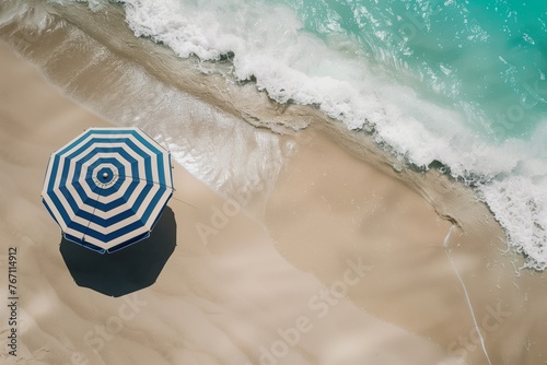 drone shot of bluestriped beach umbrella on sandy shore with turquoise waves nearby photo