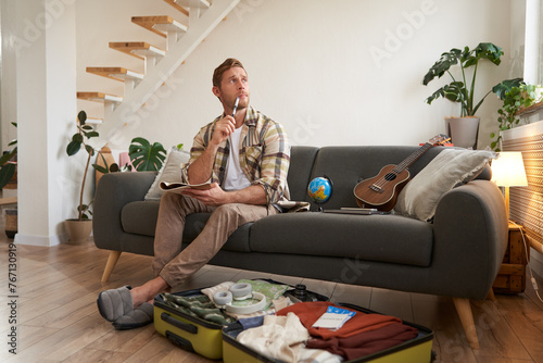 Portrait of man thinking what to take with him on vacation, holding notebook, writing list of items with thoughtful face, preparing to go on holiday, sitting near suitcase with clothes © Mix and Match Studio